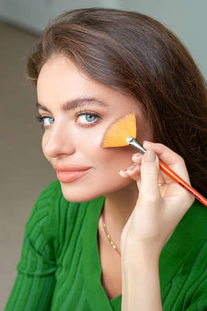 Professional make up artist applying powder by brush on cheeks of beautiful young caucasian woman in beauty salonの写真素材