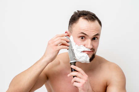 Young caucasian man shaving beard with knife on white backgroundの写真素材