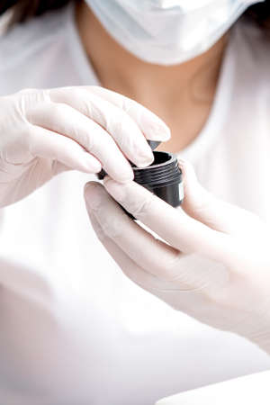 Beautician or manicure master with protective mask holds black jar with cosmetics or manicures...