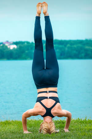 Young female doing gymnastic exercises on the grass of the lake. Woman stands on her headの写真素材