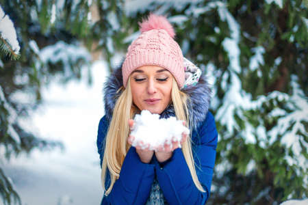 Beautiful young caucasian woman blowing on the snow holding it in her hands in winter forestの写真素材