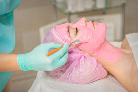 Hand of cosmetologist applying pink alginic mask to face of young woman in beauty salonの写真素材