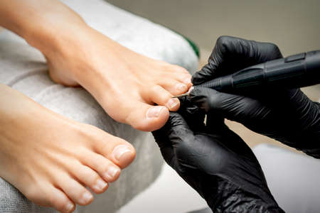Pedicure master removes cuticle from toes of woman using professional electric nail hardware in nail...