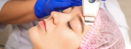Young woman receiving facial skin cleaning by ultrasonic cosmetology face equipment in medical salon