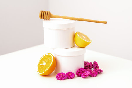 White cosmetic jars with a wooden honey stick, fresh lemon, and chrysanthemums flowers on white...