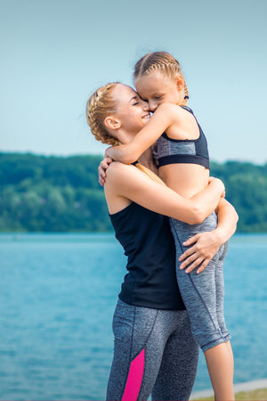 Mother embracing her daughter near the pond wearing sportswear outdoorsの写真素材