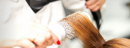 Close up of hairdresser drying long red hair with a hairdryer and round brush in a beauty salonの写真素材