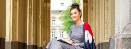 Portrait of a young female student with a book sitting on the stairs of building outdoors