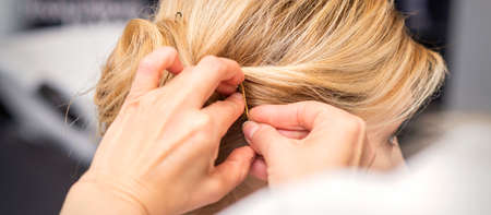 Closeup back view of a hairdresser stabs blonde hair with a hairpin for a young woman in a hair...