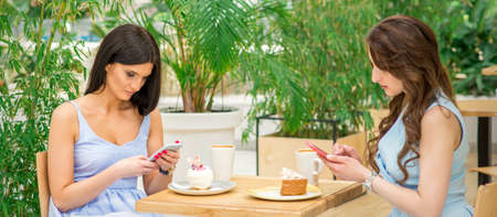 Two young women looking their smartphones while sitting in a cafe. Technology people addictions
