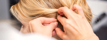 Closeup back view of a hairdresser stabs blonde hair with a hairpin for a young woman in a hair...