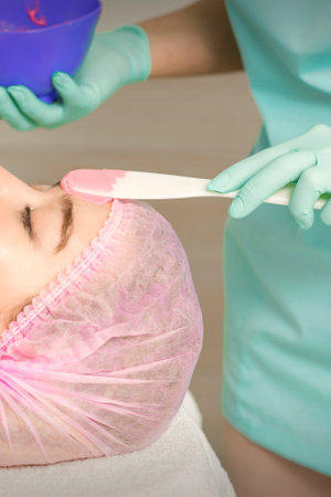 Close up of beautician's hand applying alginate mask on the face of the young caucasian woman in a...
