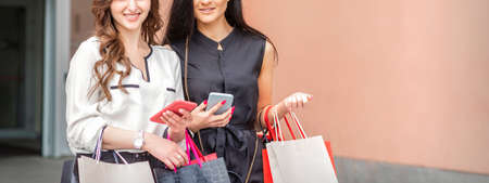 Portrait of two beautiful young caucasian women standing with paper bags and smartphones looking at the camera outdoorsの写真素材