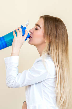 Side view of beautiful young caucasian woman is drinking water from plastic bottle against a beige backgroundの写真素材