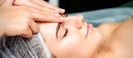 Side view fingers of female therapist making anti-age massage of forehead wrinkles to young caucasian woman in spa beauty salonの写真素材