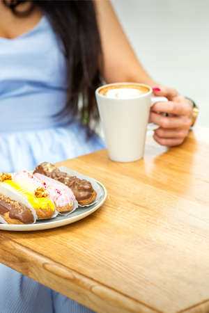 A young woman drinking coffee with sweet appetizing eclairs sitting at the table in a cafe
