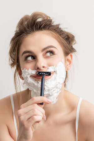 Beautiful young caucasian smiling woman shaving her face with razor looking up on white backgroundの写真素材