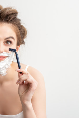 Beautiful young caucasian smiling woman shaving her face with razor looking up on white backgroundの写真素材