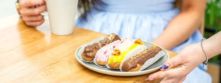 Hand of waiter puts a plate with eclairs on the wooden table for young woman drinking coffee in a...