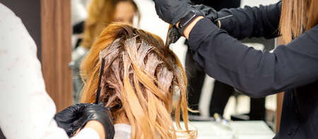 Back view of a female hairdresser in gloves coloring hair of redhead young woman with light or white dye in a beauty salonの写真素材