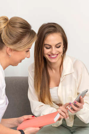 Two young women doctor with pacient smiling and looking at smartphone sitting in hospital office, consultation with a doctorの写真素材