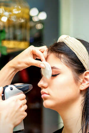Side view of a make-up artist using aerograph making an airbrush makeup foundation on a female face...