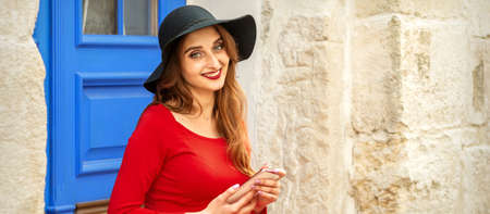 Portrait of tourist young caucasian woman in a red long dress and black hat with suitcase sitting on the stairs outdoorsの写真素材