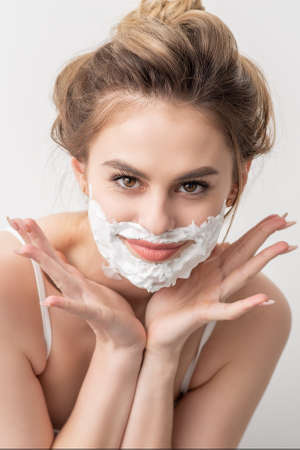 Portrait of a beautiful young smiling caucasian woman with shaving foam on face poses on white backgroundの写真素材