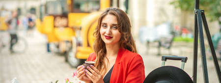 Tourist young caucasian woman in red jacket with coffee cup at the table in cafe outdoorsの写真素材