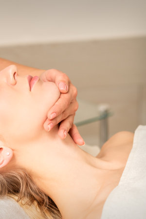 Cosmetologist hands doing facial massage on forehead and chin of young female at spa salonの写真素材