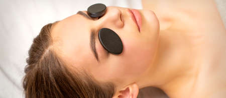 Black massage stones lying on the eyes of the young caucasian woman. Facial massage in a spaの写真素材
