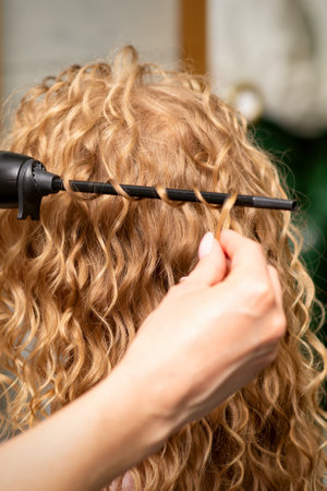 Hands of hairstylist curl wavy hair of young woman using a curling iron for hair curls in the beauty salon rear viewの写真素材