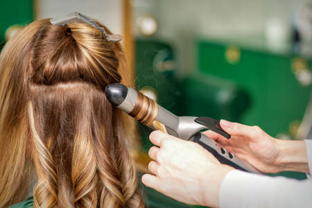 Creating curls with curling irons. Hairdresser makes a hairstyle for a young woman with long red...