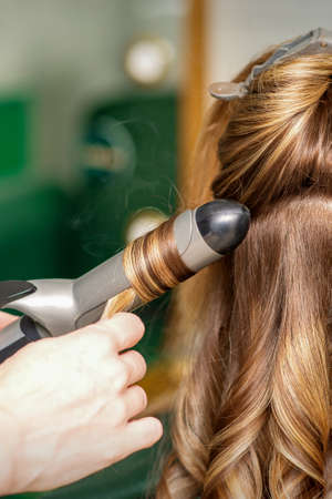 Creating curls with curling irons. Hairdresser makes a hairstyle for a young woman with long red hair in a beauty salonの写真素材