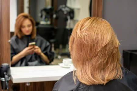 Beautiful young red hair woman using her smartphone and texting sitting in front of a mirror waiting to visit a hairdresserの写真素材