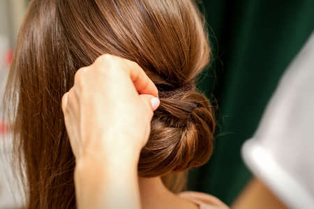 Hands of hairdresser making french twist hairstyle of an unrecognizable young brunette woman in a...