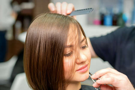 The hairdresser cuts the hair of a brunette woman. Hairstylist is cutting the hair of female client in a professional hair salon, close upの写真素材
