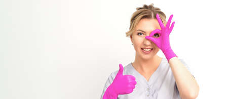 Smiling doctor oculist caucasian woman wearing pink rubber gloves in uniform showing ok sign covering the eye and thumb up gesture against a white backgroundの写真素材