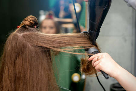 A hairdresser is drying long brown hair with a hairdryer and round brush in a hairdressing salonの写真素材