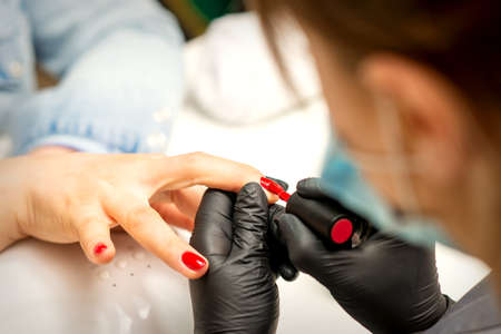 Manicure varnish painting. Close-up of a manicure master wearing rubber black gloves applying red varnish on a female fingernail in the beauty salonの写真素材