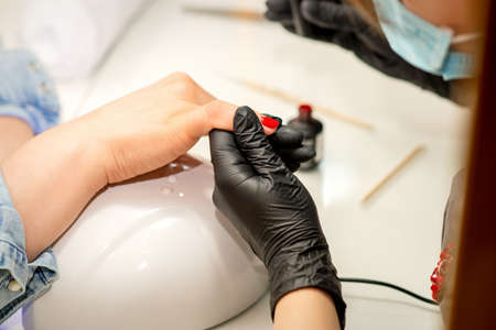 Manicure varnish painting. Close-up of a manicure master wearing rubber black gloves and a protective mask applying red varnish on a female fingernail in the beauty salonの写真素材