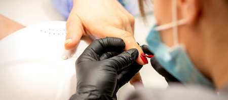 Manicure varnish painting. Close-up of a manicure master wearing rubber black gloves applying red...