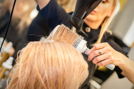 Drying straight blond hair with black hairdryer and white round brush in hairdresser salon, close upの写真素材