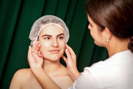 Plastic surgery and beauty concept. Professional cosmetician examining and touching the face of the beautiful young caucasian smiling woman in a clinic of esthetic cosmetologyの写真素材