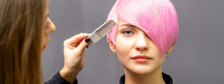 The female hairdresser is combing the dyed pink short hair of the young woman against the dark wallの写真素材