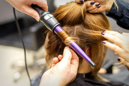 The hairstylist makes curls hairstyle of long brown hair with the curling iron in hairdresser salon, close upの写真素材