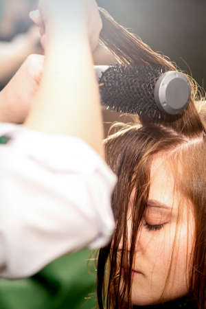 A hairdresser is drying long brown hair with a hairdryer and round brush in a beauty salonの写真素材