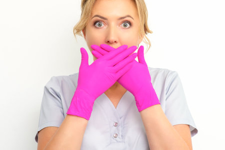 Portrait of a young female caucasian doctor or nurse is shocked covering her mouth with her pink gloved hands against a white backgroundの写真素材