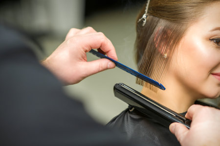 Hairstylist is straightening short hair of young brunette woman with a flat iron in a hairdresser salon, close upの写真素材