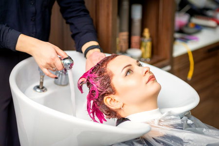 Washing dyed female hair. A young caucasian woman having her hair washed in a beauty salon. Professional hairdresser washes pink color paint off of a customers hairの写真素材